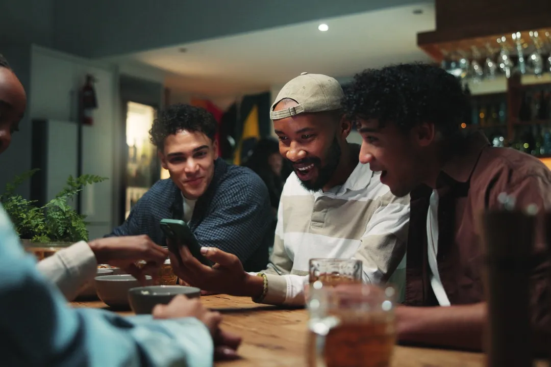 Drei Freunde sitzen zusammen am Tisch und schauen lachend auf ein Smartphone